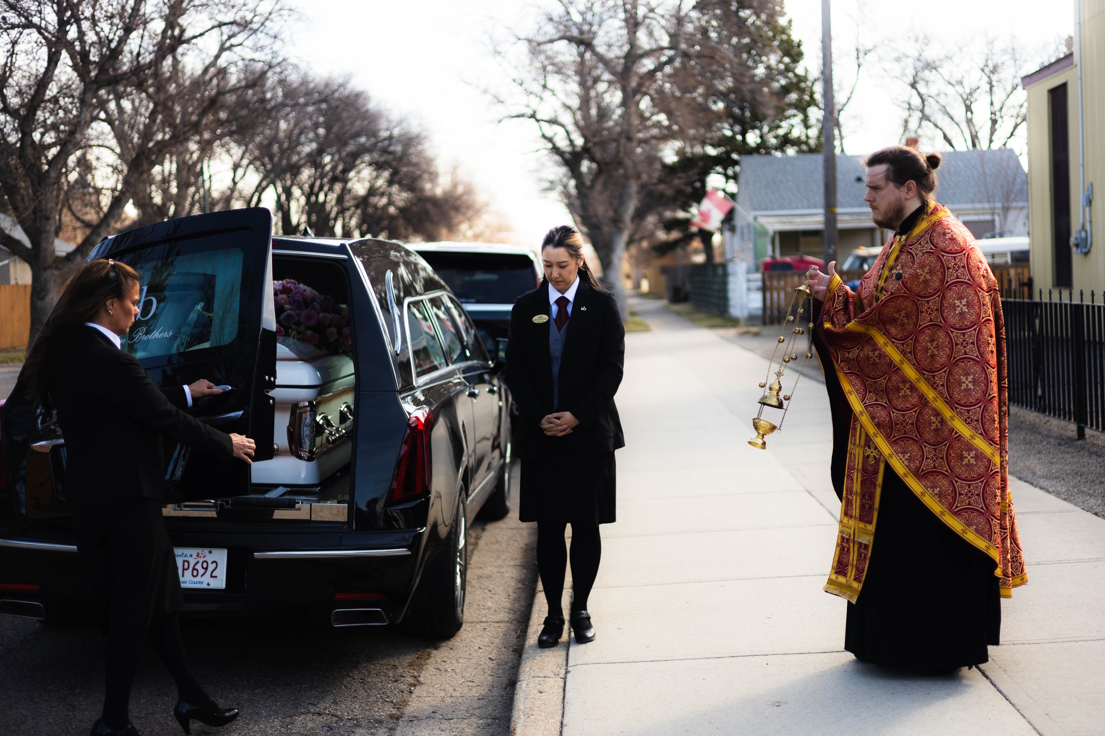 A wide-angle cinematic photograph of a formal funeral procession. A priest in ornate gold and red vestments swings a thurible on the sidewalk, while two Martin Brothers professionals in formal black attire stand in silent stewardship by the open door of a black hearse containing a white casket.