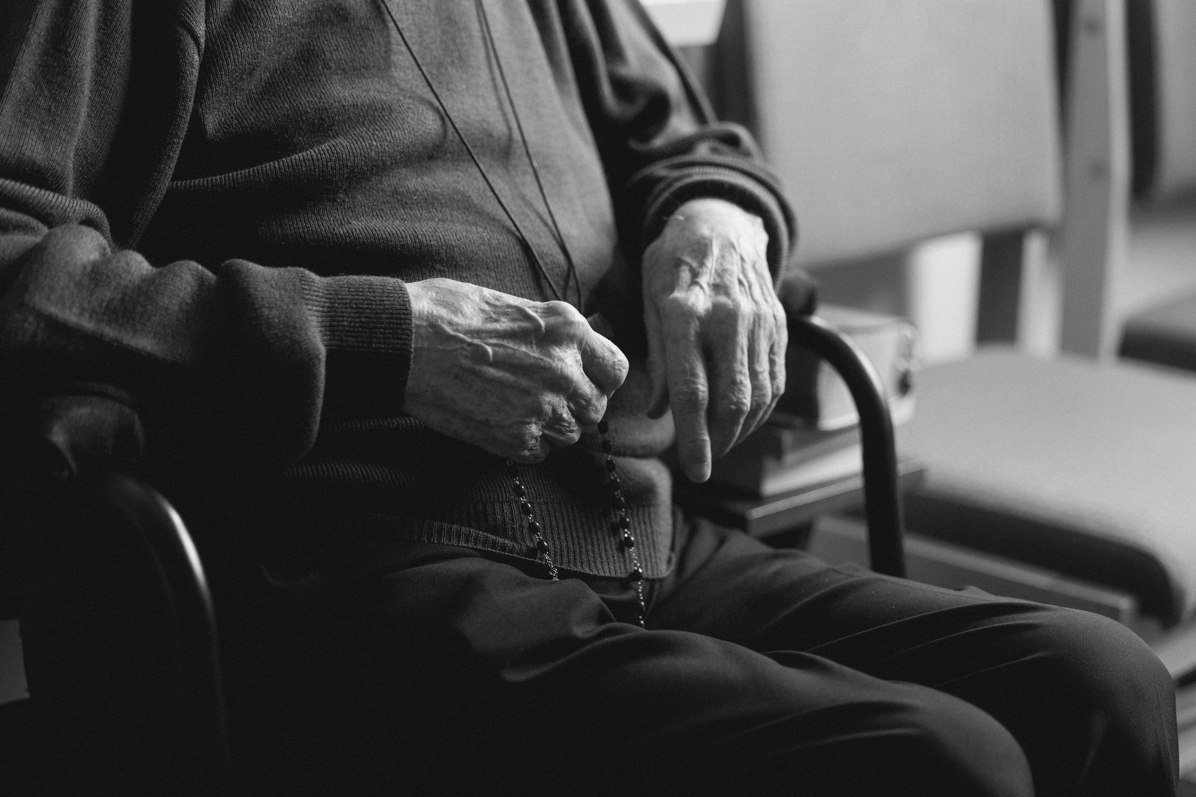 A close-up, black-and-white documentary photograph focusing on the weathered hands of an elderly person seated in a chair, quietly holding a beaded rosary in their lap. In the context of this case study, this image acts as a study in visual literacy, using the contrast of light and texture to communicate a profound narrative of stillness, faith, and lived experience without a single word.